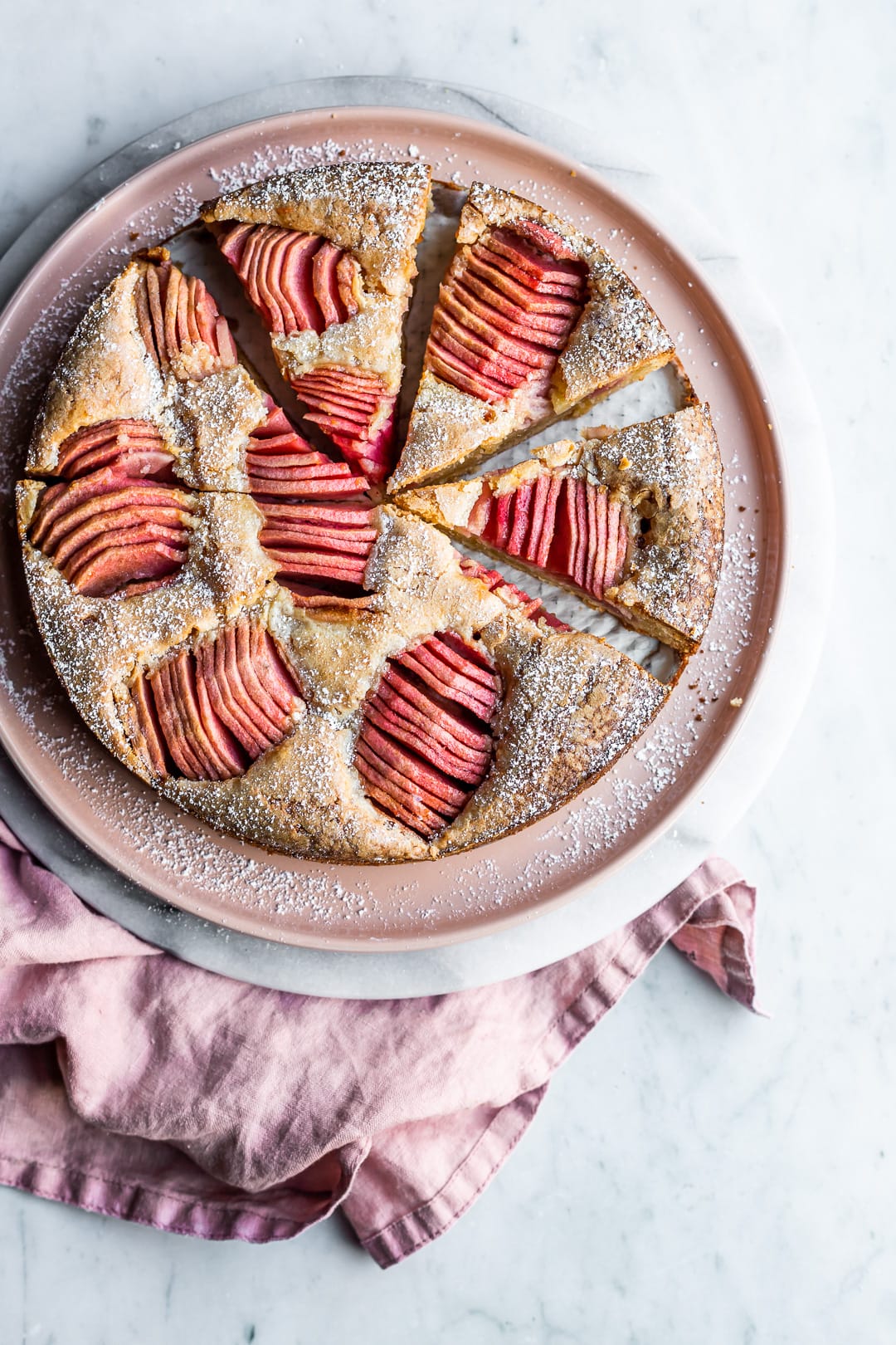 Sliced almond cake with pink apples on pink plate and marble surface