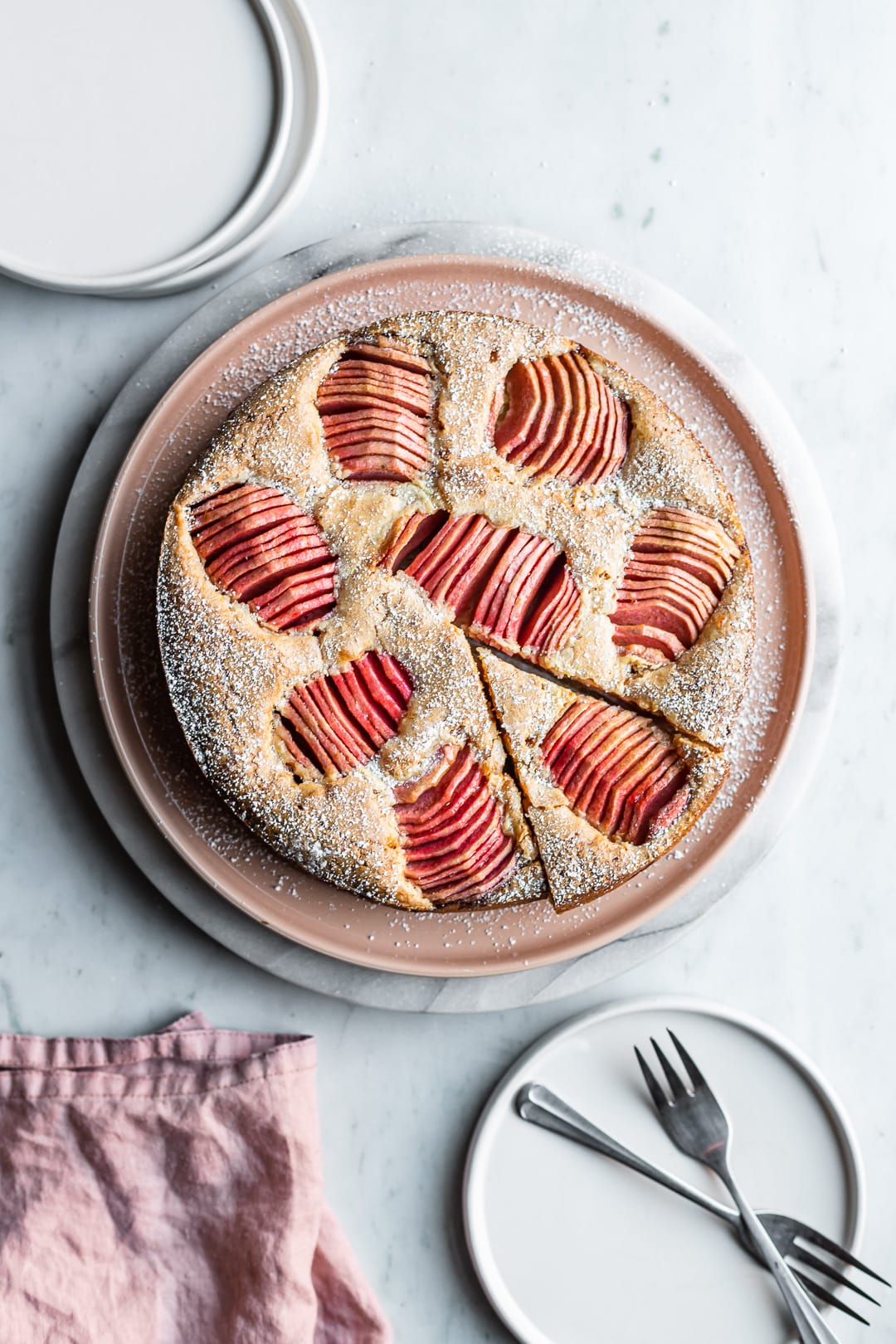 Cake with apples with a slice cut out, on a white marble background and white plates nearby