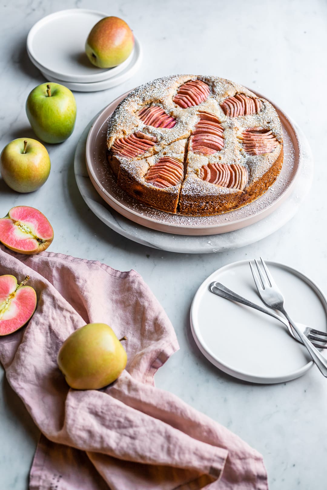 Cake on plate surrounded by pink apples and white plates on a marble background