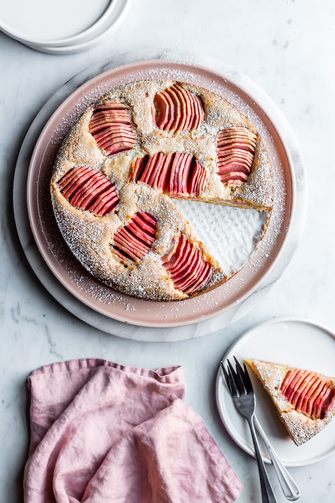 Cake with pink apples with a slice cut out on a plate nearby on a grey marble background