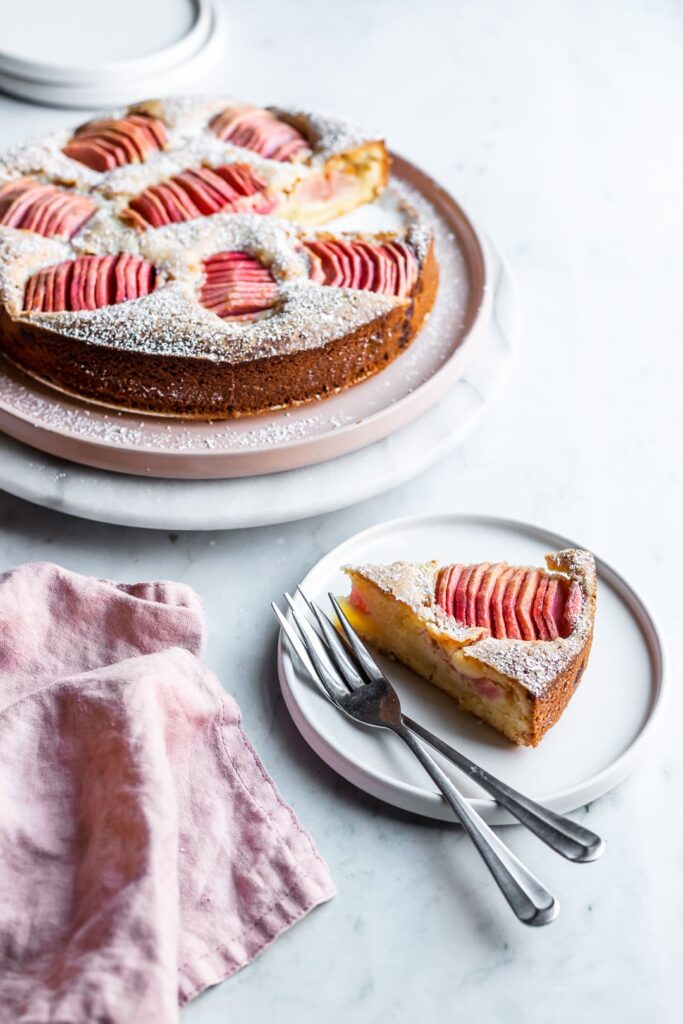 A slice of almond cake with pink apples with the rest of the cake nearby. 