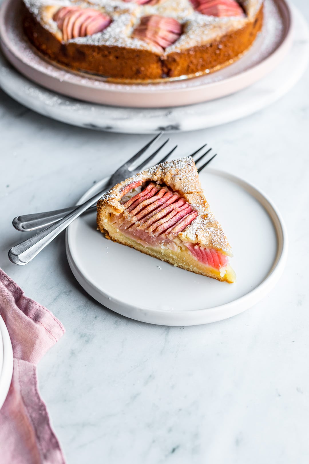 Slice of almond cake with pink apples on a white plate and marble background