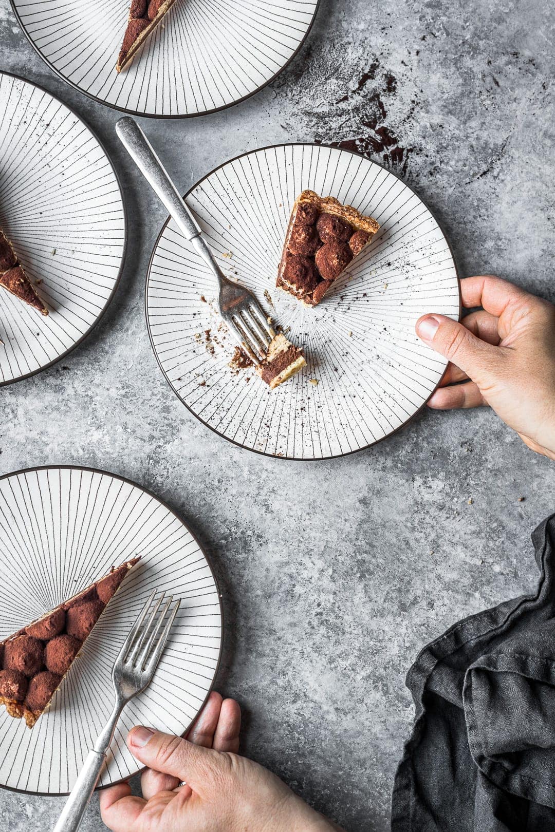 Hands holding plates with slices of tart and forks, all on a grey background