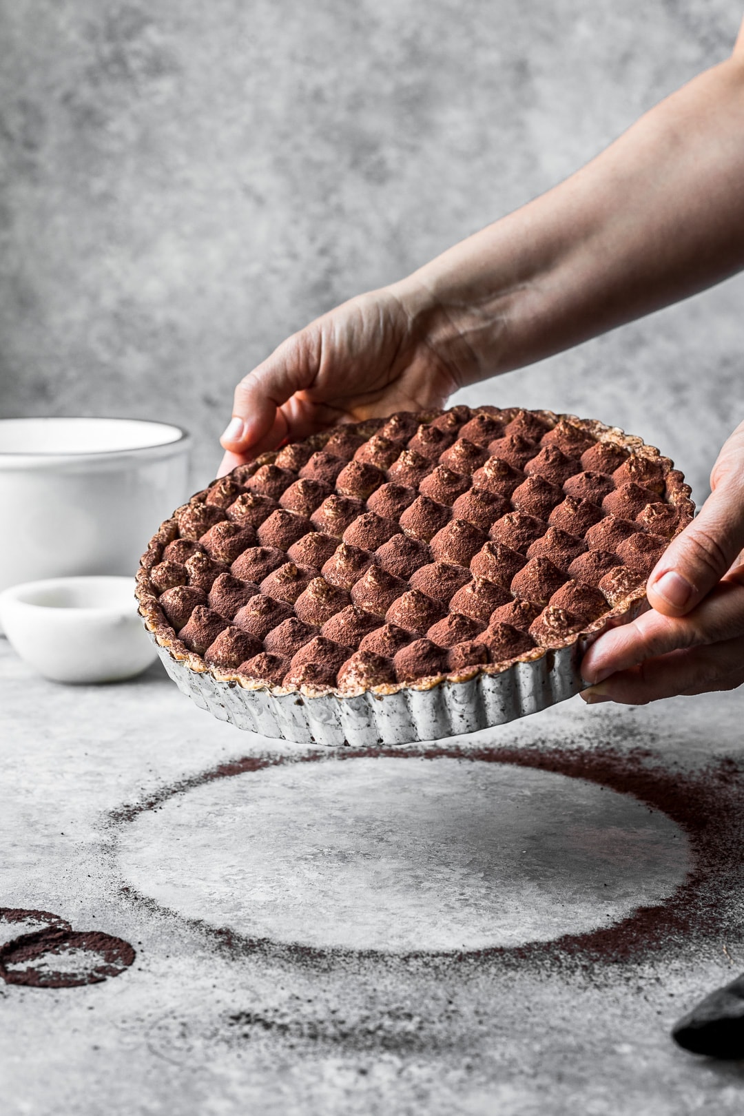 Hands holding a chocolate coffee mascarpone tart for display against a grey background