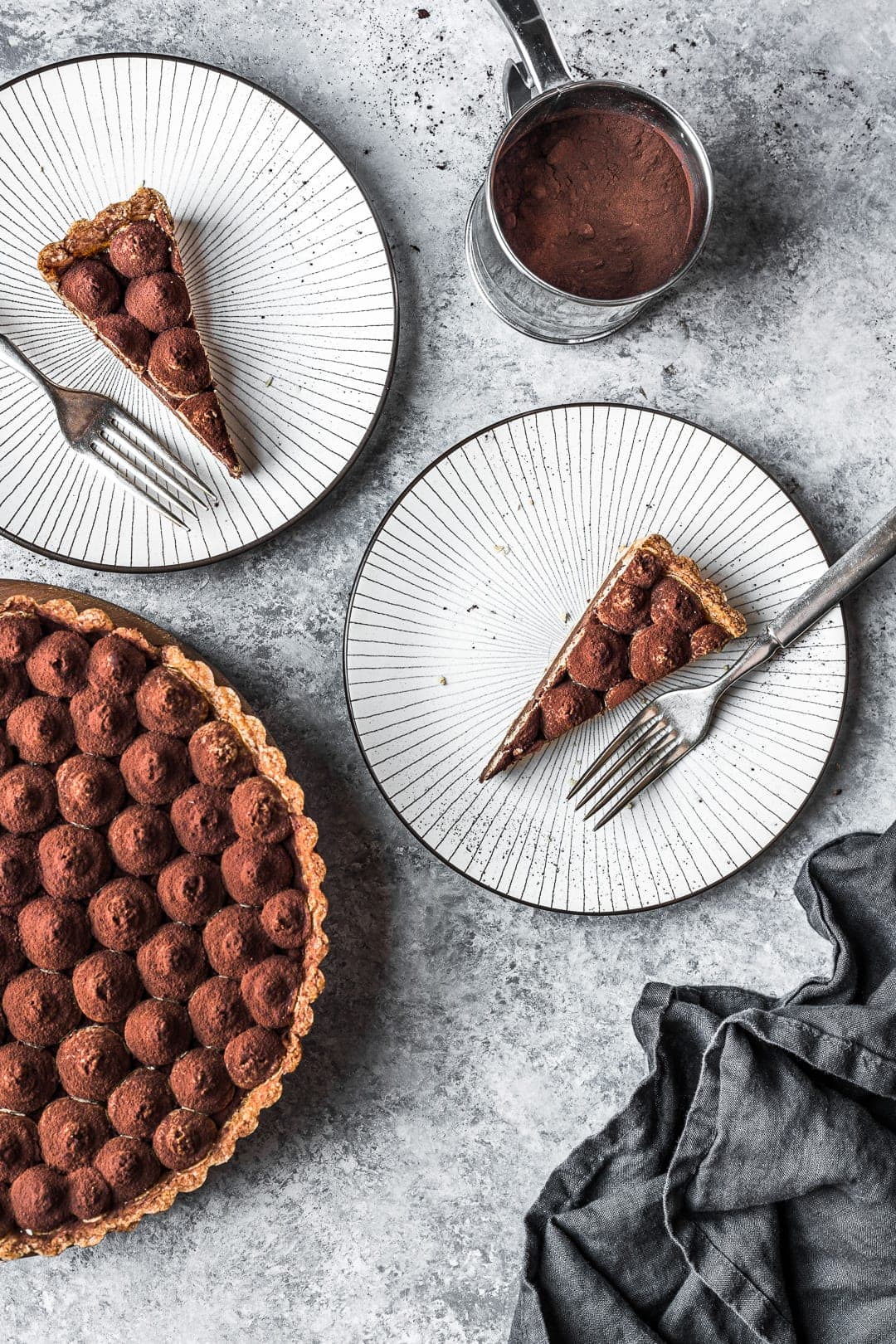 Two slices of tart on plates with rest of tart and sifter of cocoa powder nearby
