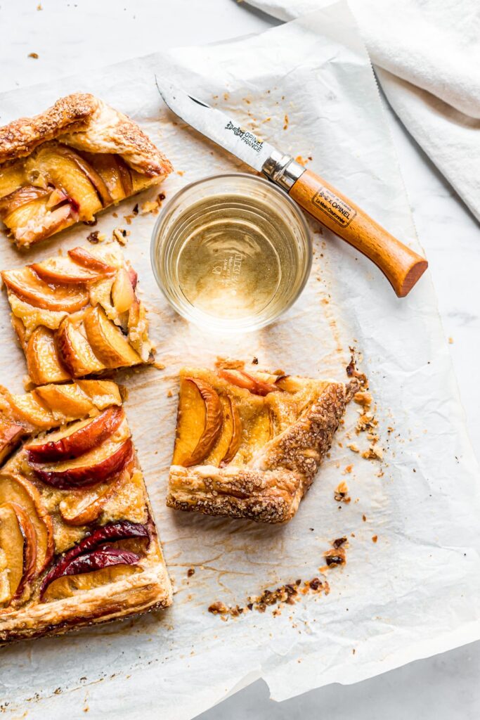Peach galette slices on parchment paper and a cutting board with white wine and knife nearby