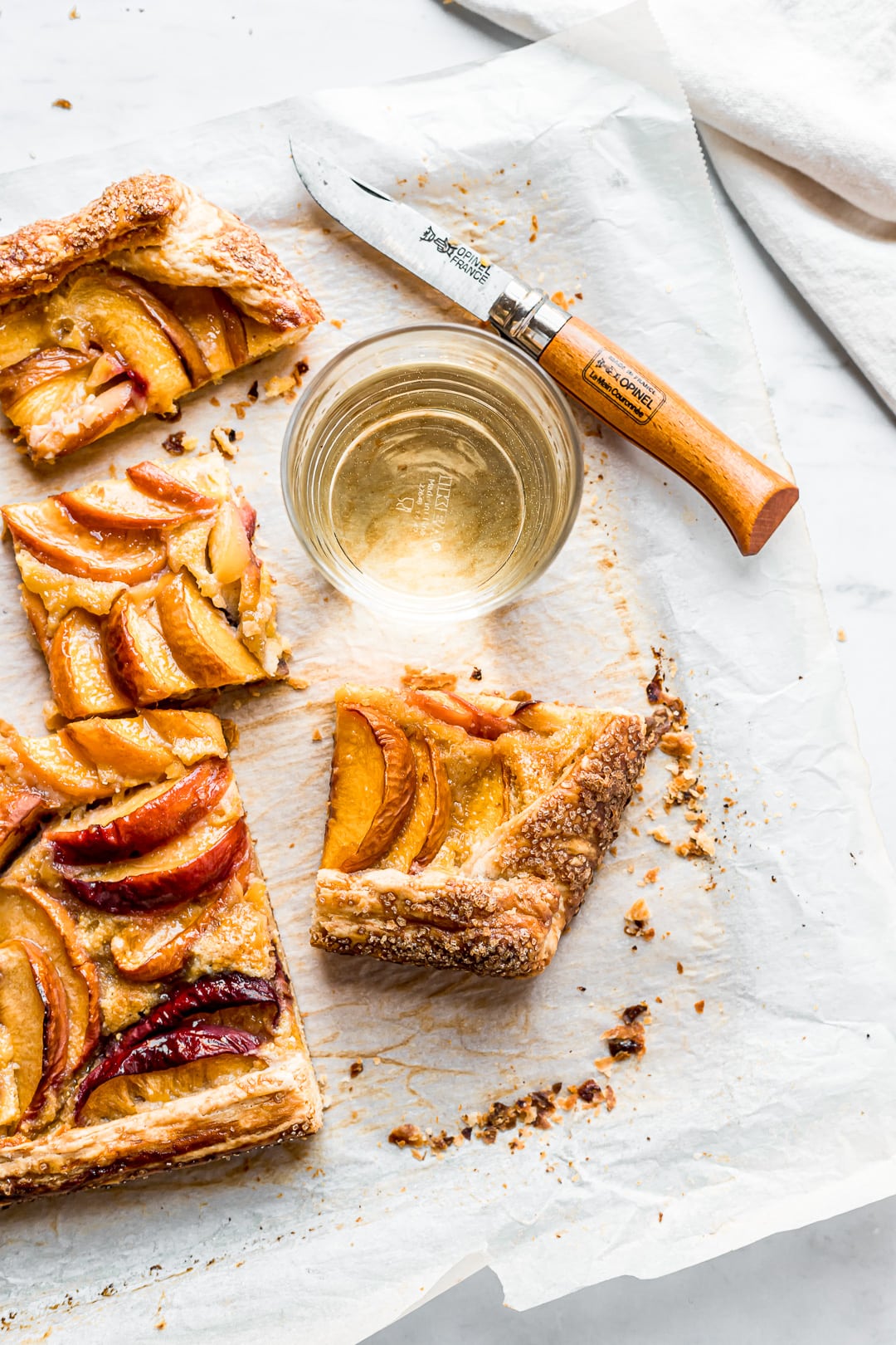 Peach galette slices on parchment paper and a cutting board with white wine and knife nearby