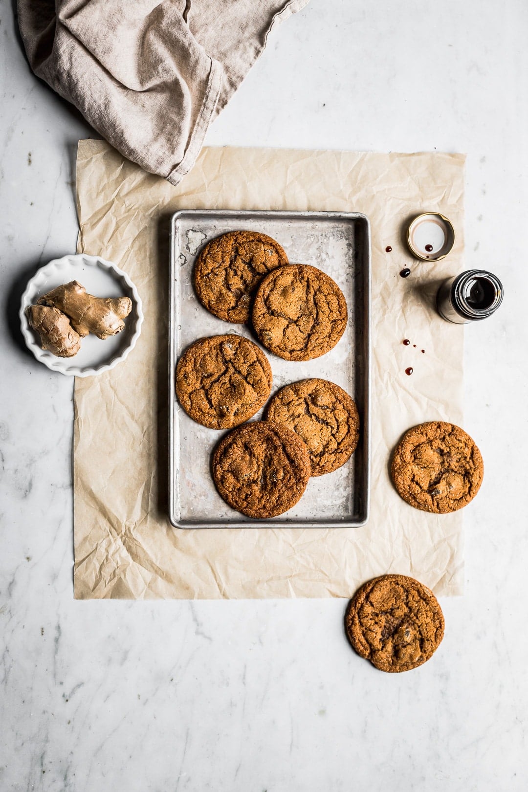 Top view of cookies on small baking sheet with ginger and molasses nearby on white marble background