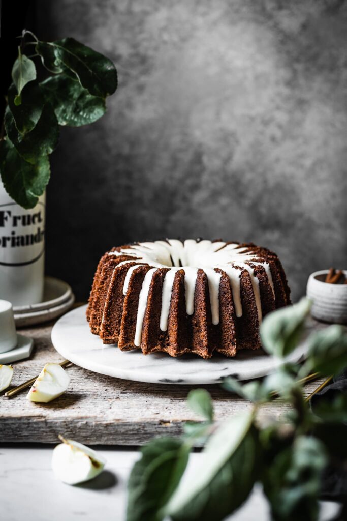 Brown bundt cake with white glaze on a marble platter with apple branches in foreground