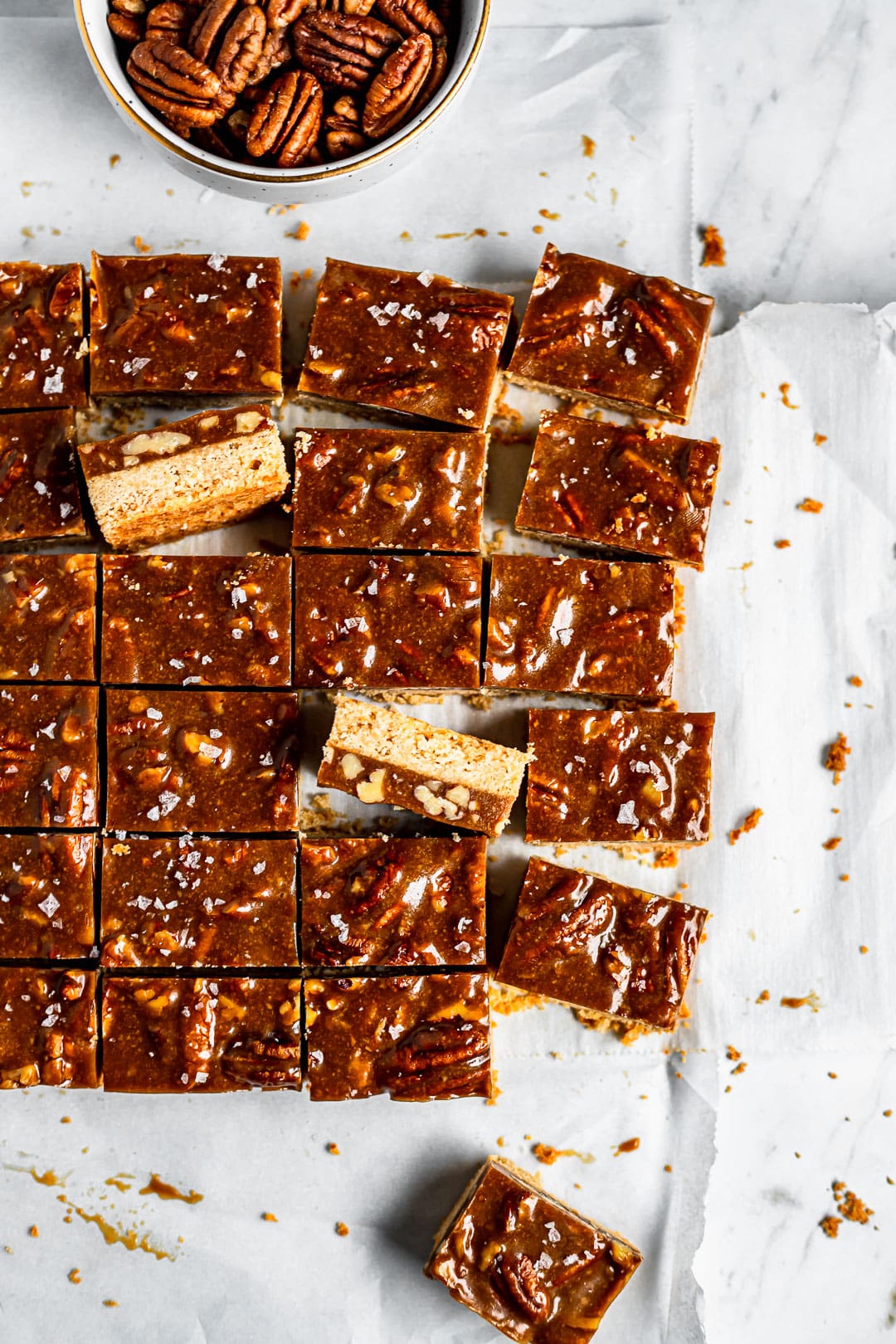 Slices of pecan bars on white parchment paper with a bowl of pecans nearby
