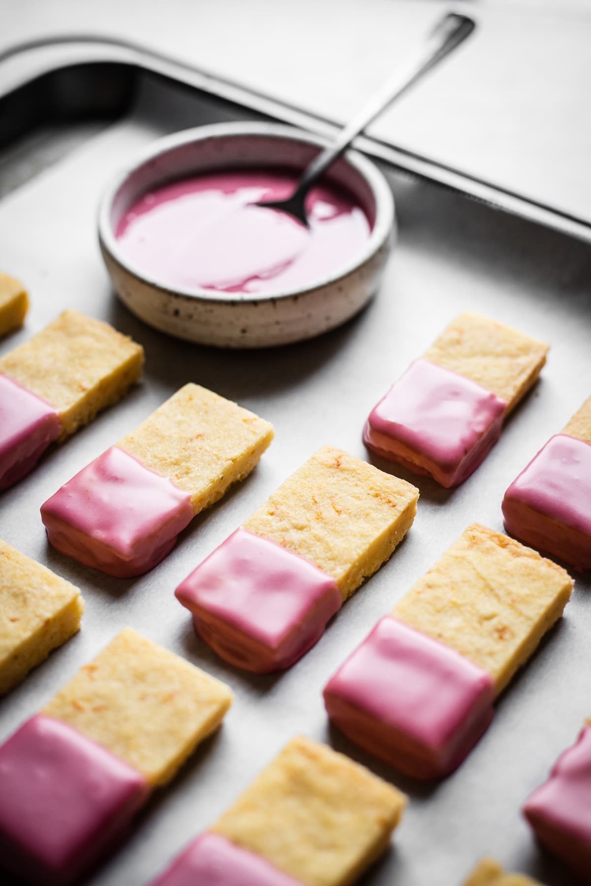 Shortbread bars dipped in pink glaze drying on a parchment lined cookie sheet with a bowl of glaze nearby