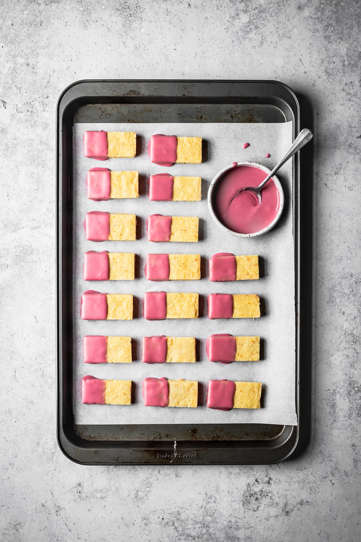 Top view of rows of yellow cornmeal shortbread cookies dipped in pink glaze drying on a parchment lined cookie sheet