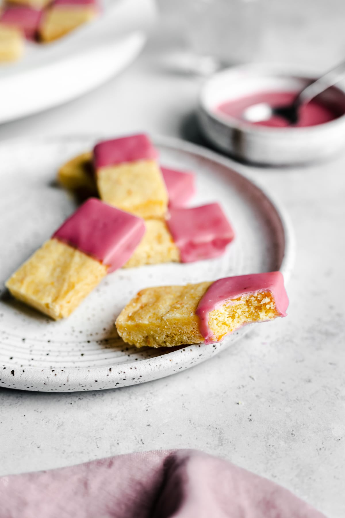 Pink glazed shortbread cookies on a white plate with a bowl of glaze in background