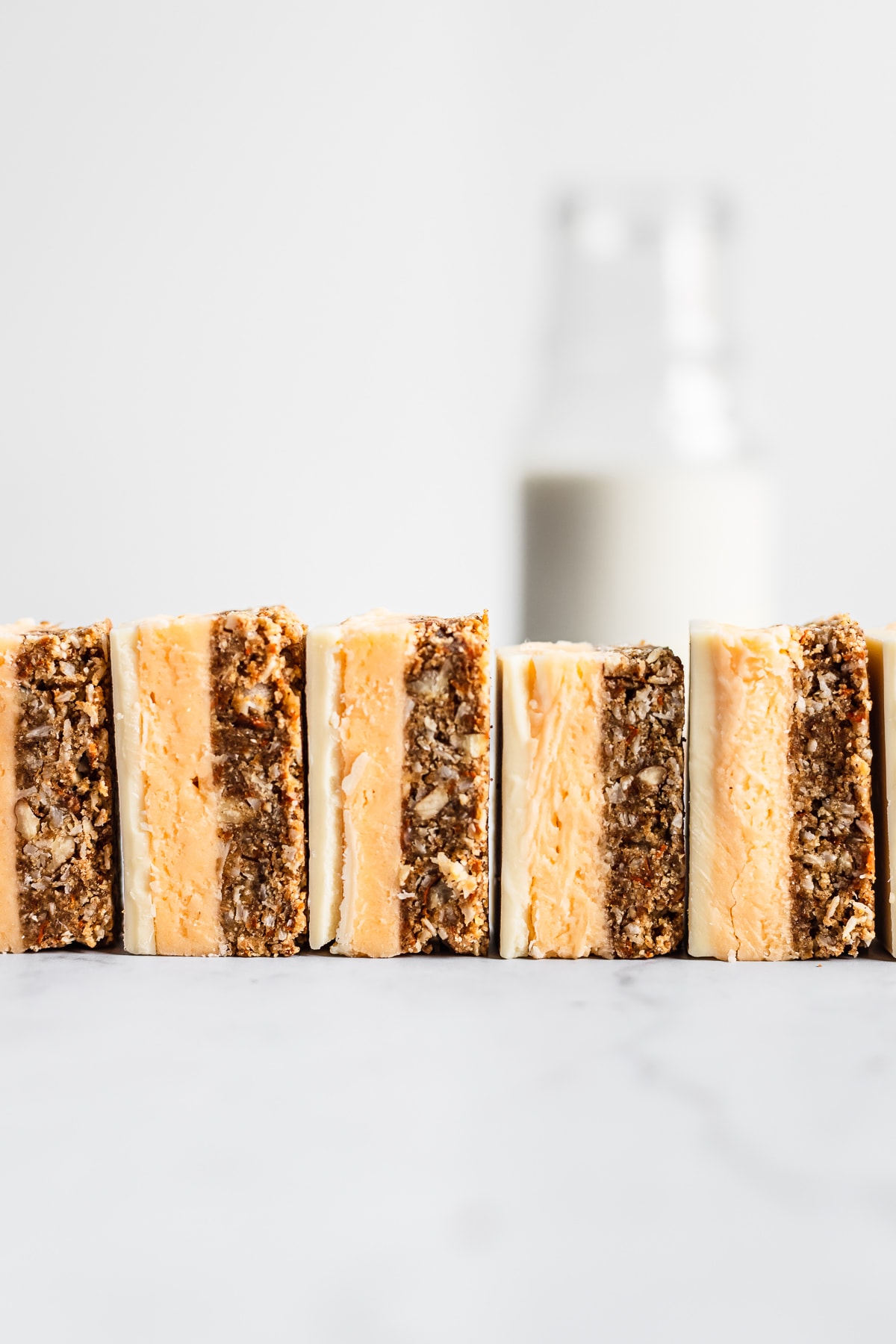 A row of sliced carrot cake bars turned on their side and lined up like dominos on a white marble surface with a glass bottle of milk in the background