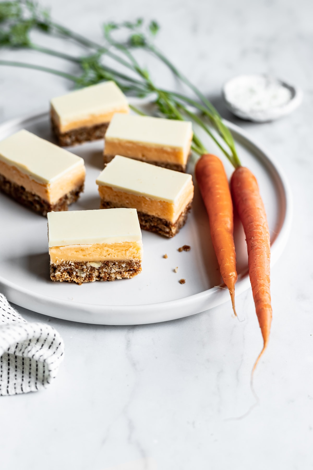 Sliced carrot cake bars on a white plate surrounded by carrots, a bowl of coconut on a white marble background