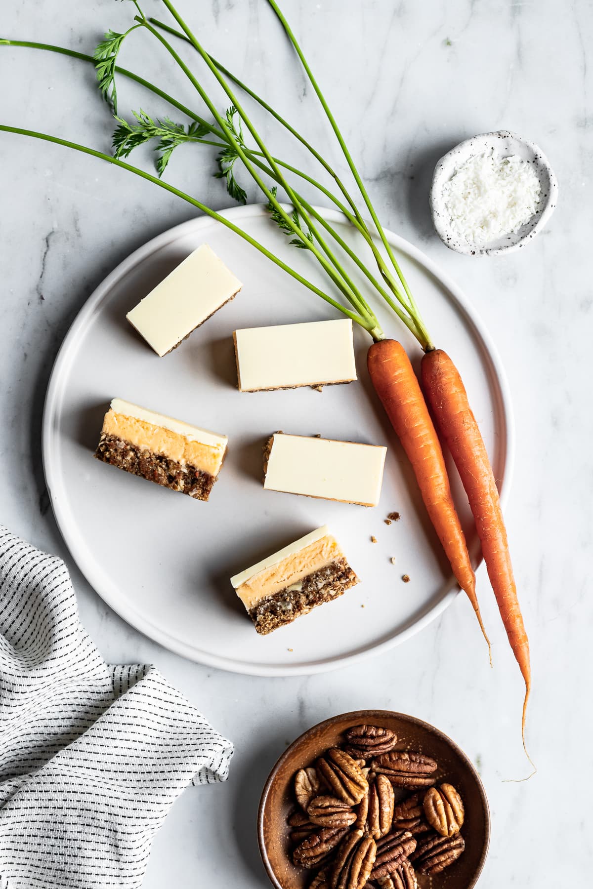 Sliced bar cookies on a white plate and marble background surrounded by carrots, a bowl of coconut and a bowl of pecans on a white marble background