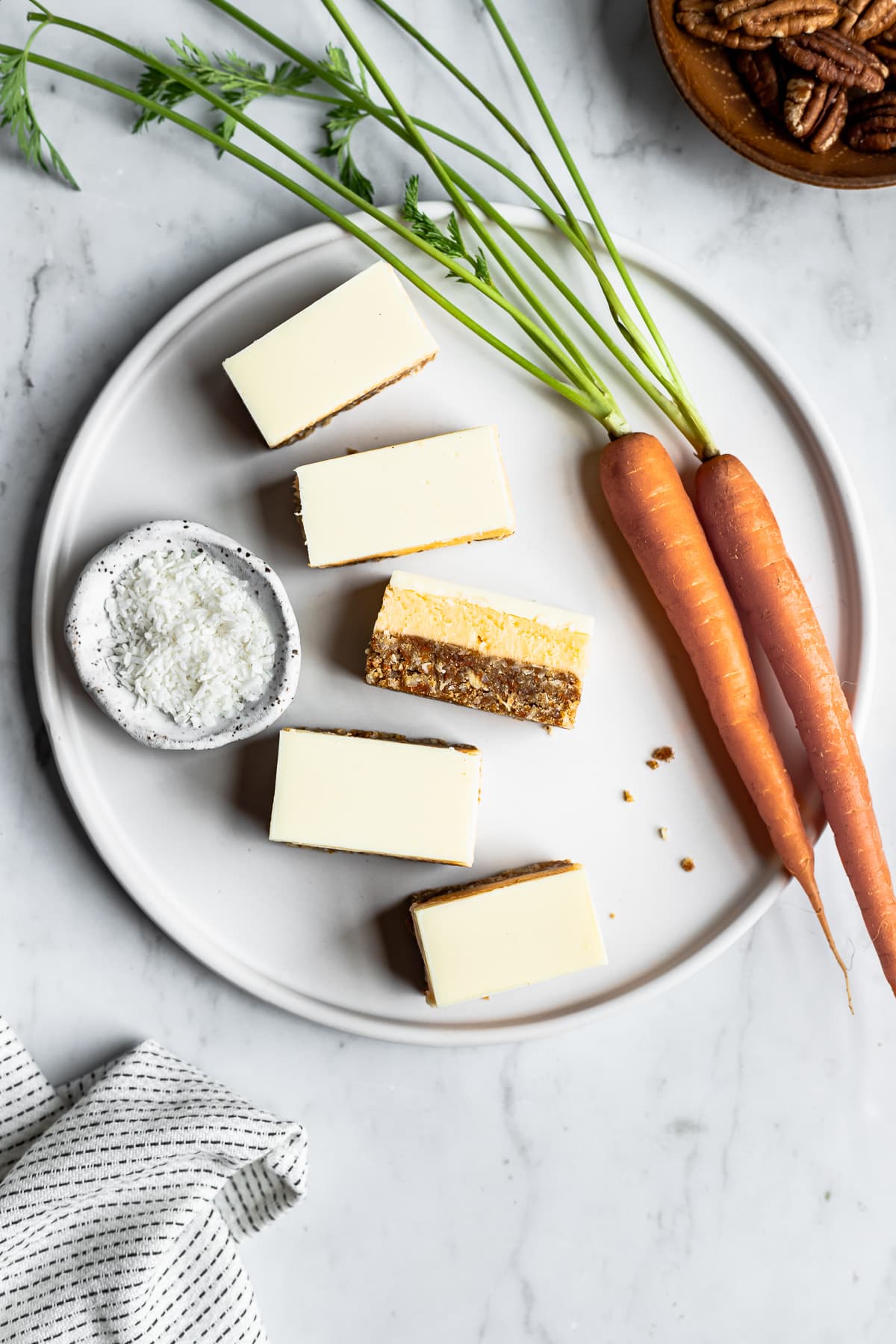 Sliced bar cookies on a white plate surrounded by carrots, a bowl of coconut and a bowl of pecans on a white marble background