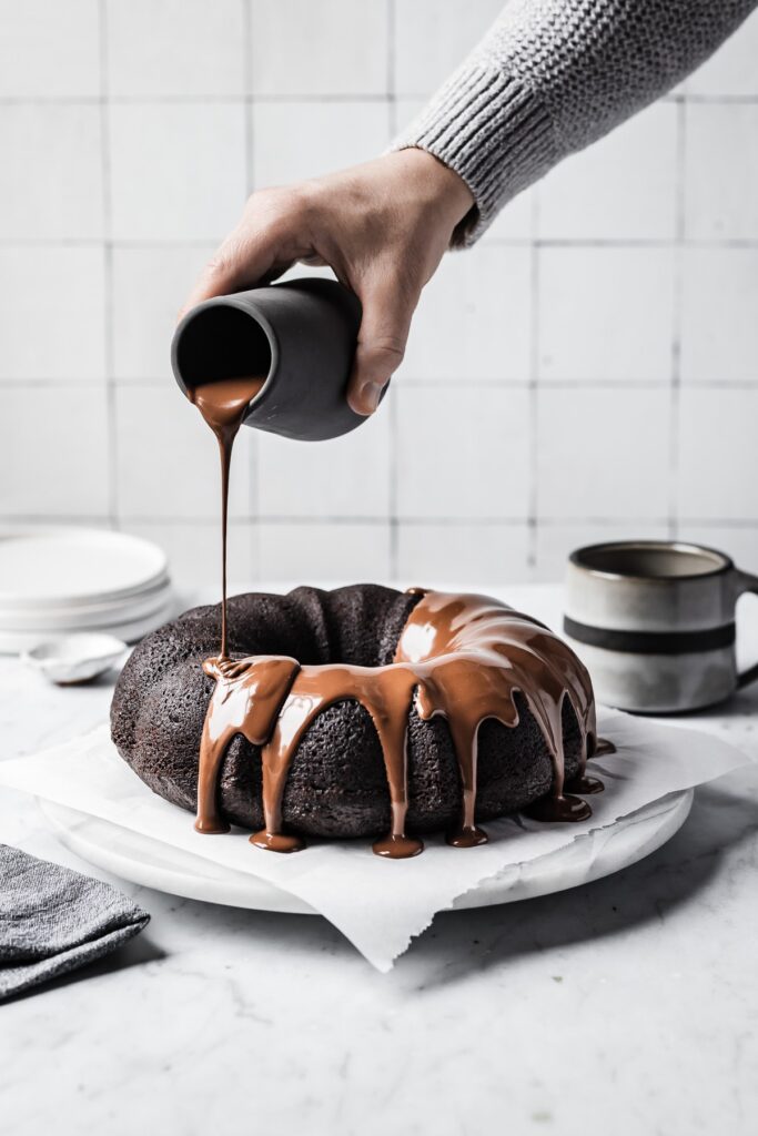 A hand pours a pitcher of chocolate ganache onto a chocolate bundt cake on a white marble surface.