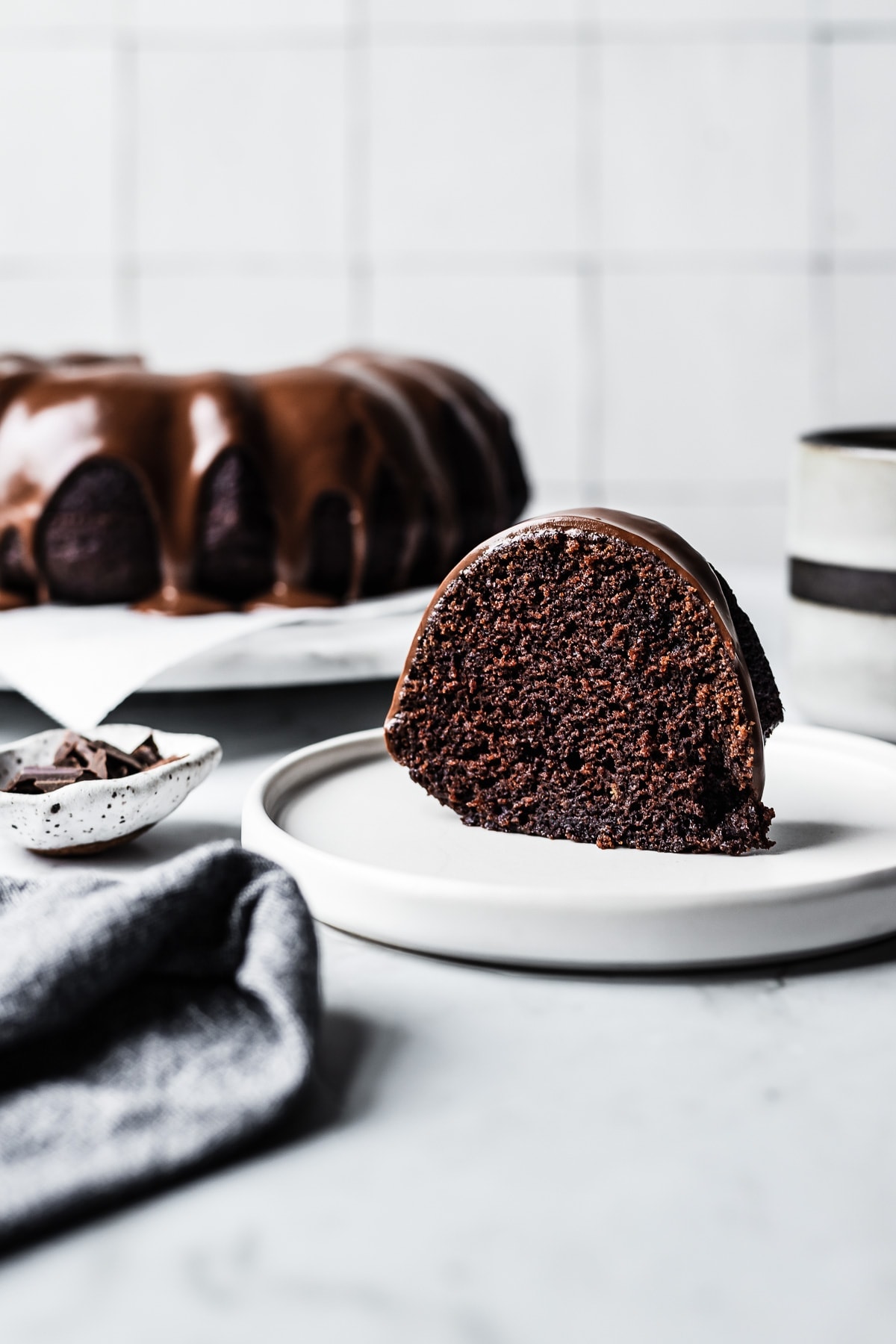 Slice of chocolate olive oil bundt cake on a white plate and marble counter with whole cake on platter in the background