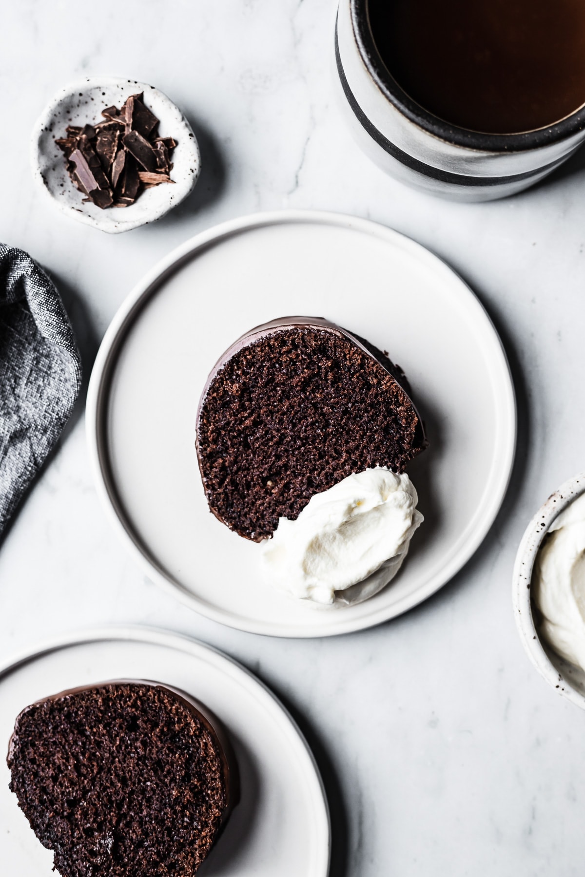 Slices of chocolate bundt cake on white plates with coffee, whipped cream and napkin resting nearby