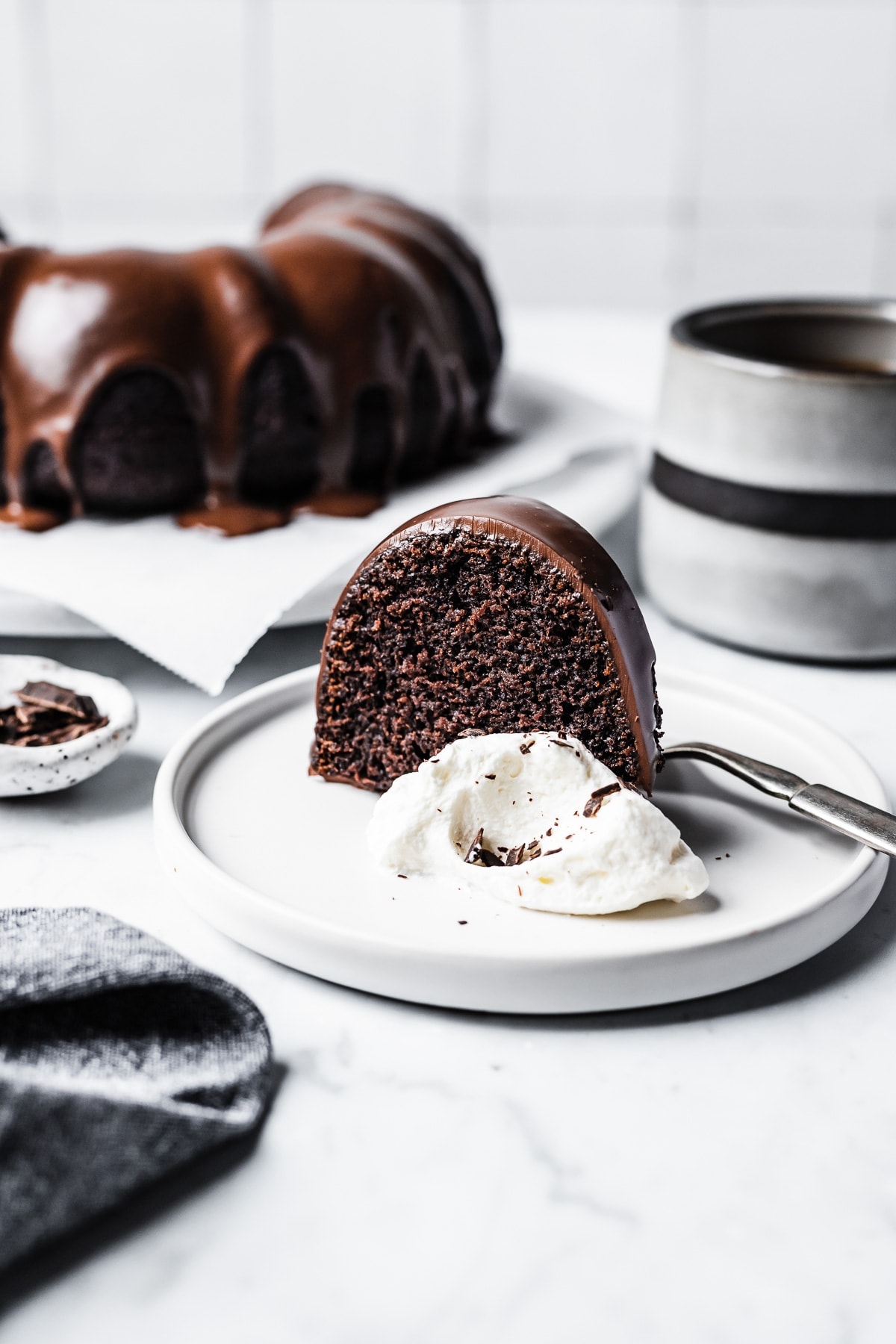 Slice of chocolate olive oil bundt cake with chocolate ganache glaze on a white plate with coffee mug and cake in background