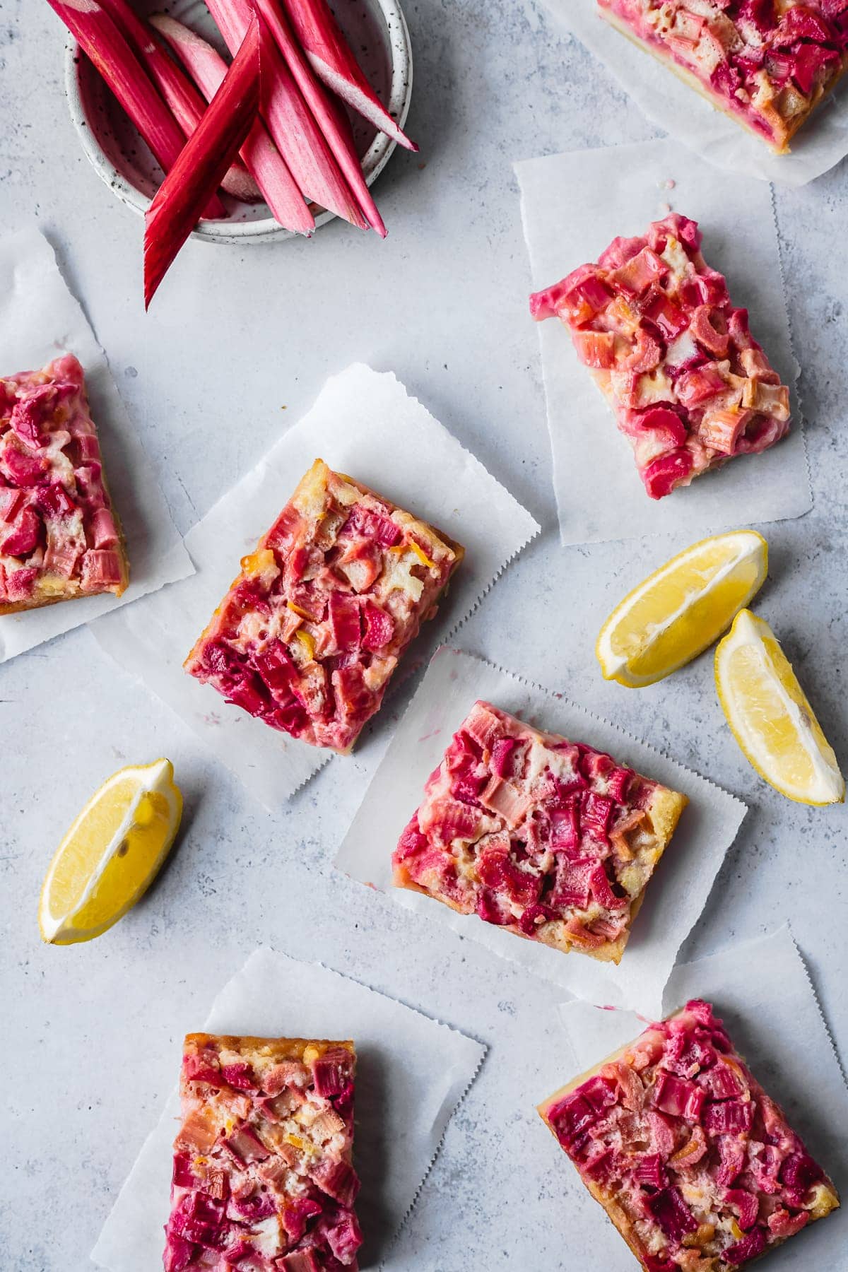 Bar cookies arranged on parchment paper strips on a white background with sliced lemons and a bowl of rhubarb nearby