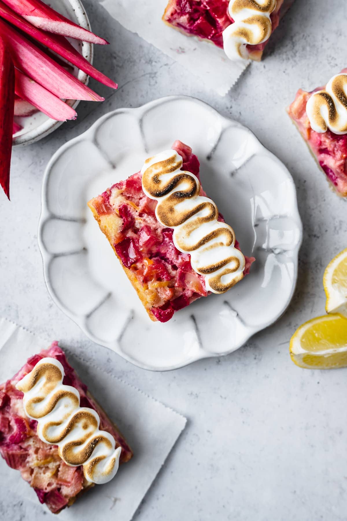 Lemon rhubarb bar on a white plate and white cement surface with lemons and rhubarb nearby