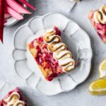 Lemon rhubarb bar on a white plate and white cement surface with lemons and rhubarb nearby