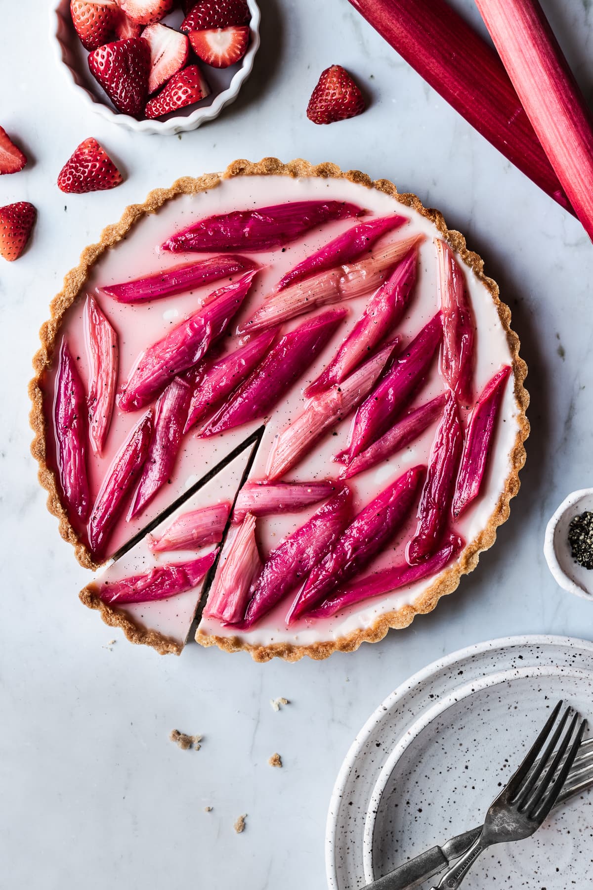 Strawberry rhubarb panna cotta tart with a slice cut into it on a grey marble background with strawberries, rhubarb and plates nearby