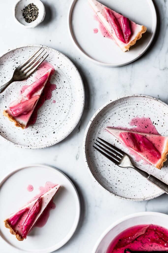 Four slices of tart topped with poached rhubarb on white plates and a grey marble background