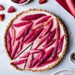 Strawberry rhubarb panna cotta tart with a slice cut into it on a grey marble background with strawberries, rhubarb and plates nearby