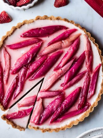 Strawberry rhubarb panna cotta tart with a slice cut into it on a grey marble background with strawberries, rhubarb and plates nearby