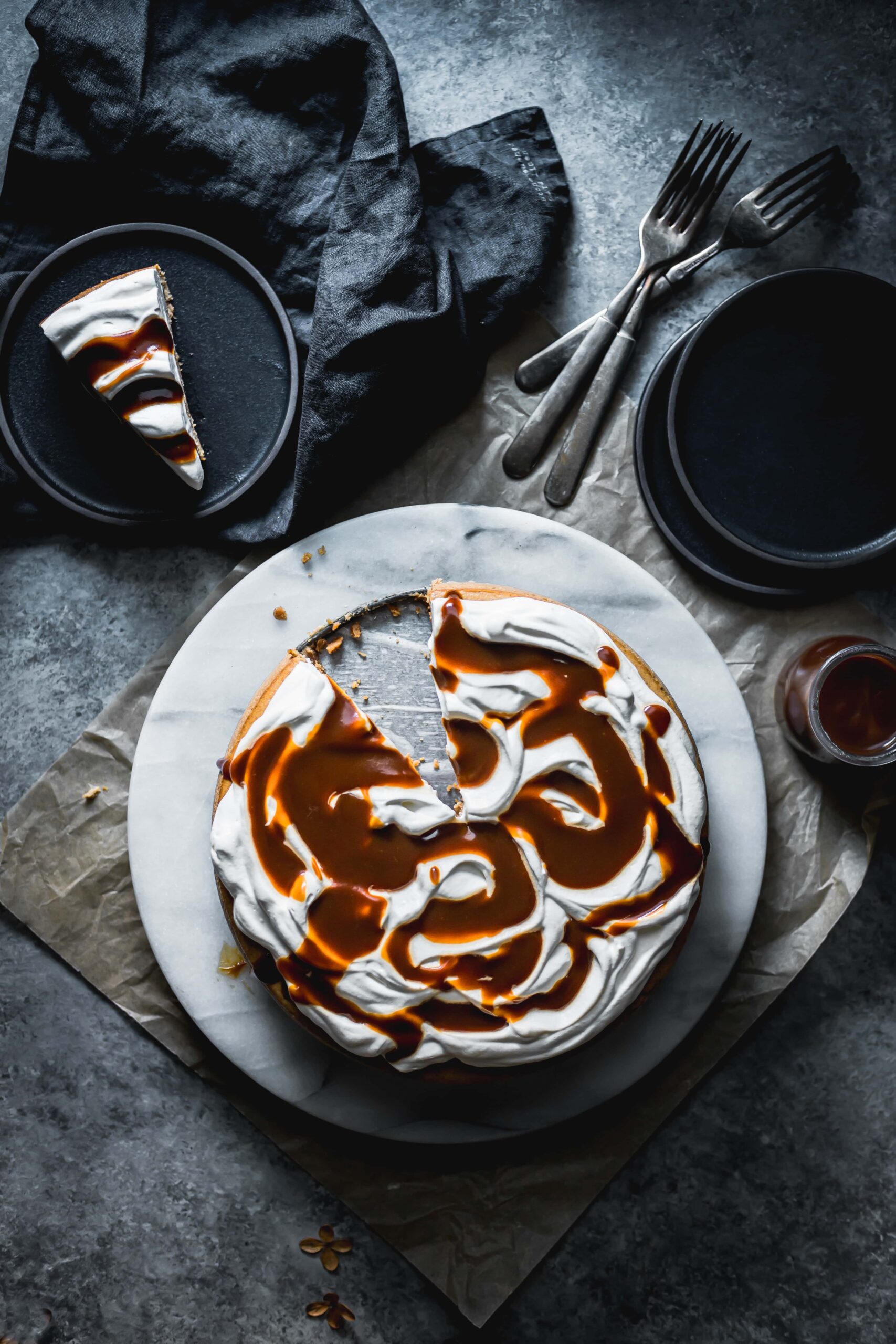 A gingerbread cheesecake topped with whipped cream and golden caramel sauce drizzled in swirls on top. The cake rests on a circular white marble platter. There is a slice cut out of the cake, and the slice sits on a black plate nearby with a black napkin, three forks, and extra black plates. The background is a dark grey concrete color, and the picture is dark and moody. 