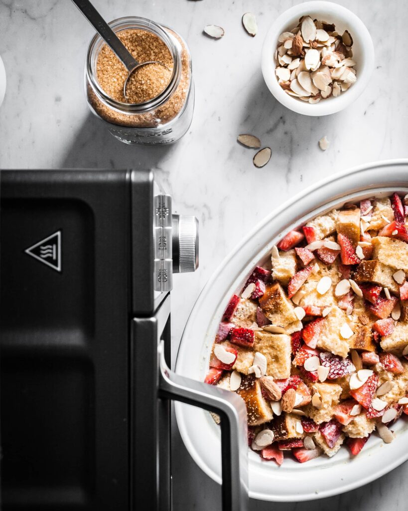 Top view of a baked strawberry french toast casserole, sprinkled with turbinado sugar and slivered almonds. A countertop oven sits nearby along with a jar of sugar and a bowl of almonds. 