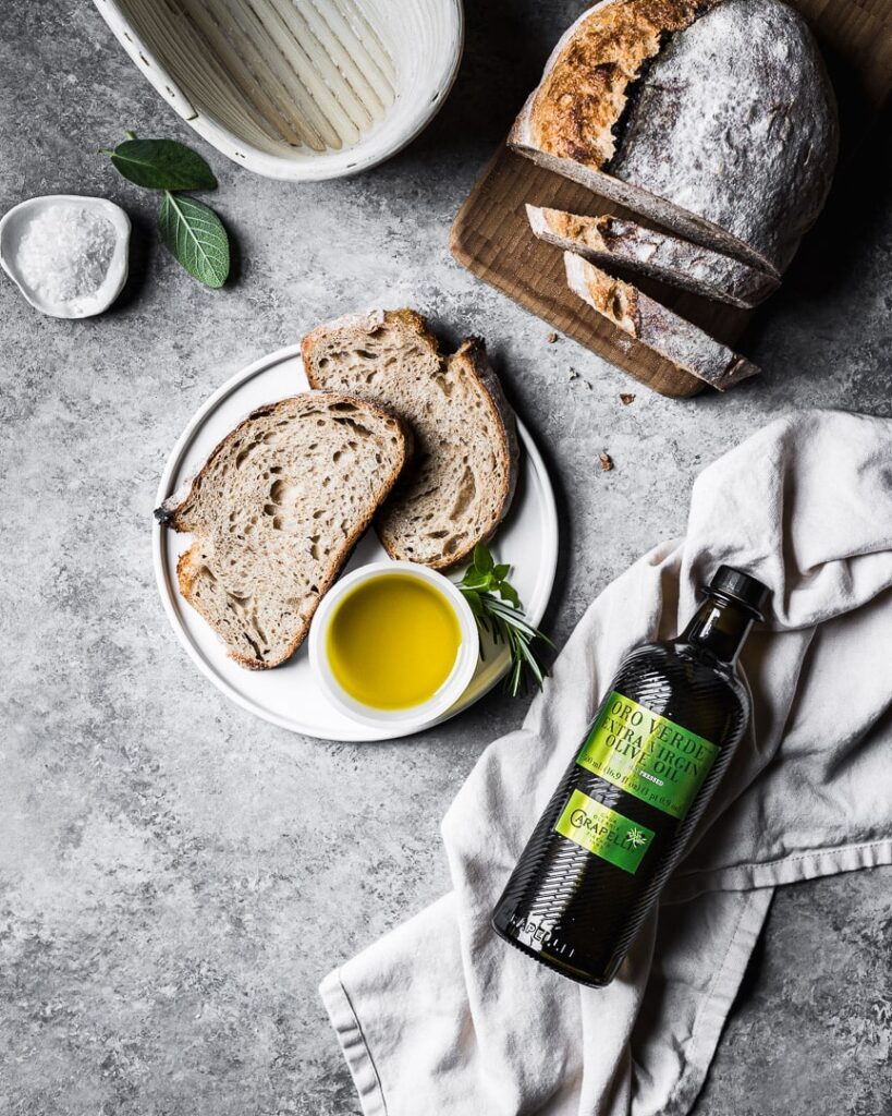 A white plate holds two sourdough bread slices with a small bowl of olive oil and herbs. Nearby, there is a bottle of olive oil with a green label, a linen napkin, a cutting board with the loaf of bread, and a bread proving basket. 