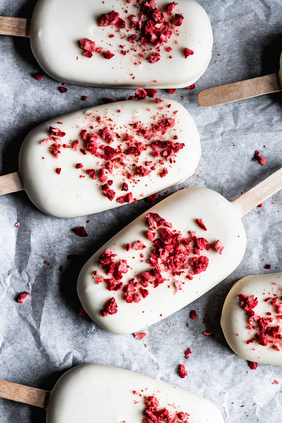 Several white chocolate covered ice cream bars with red freeze dried strawberry bits rest on a white parchment paper surface.