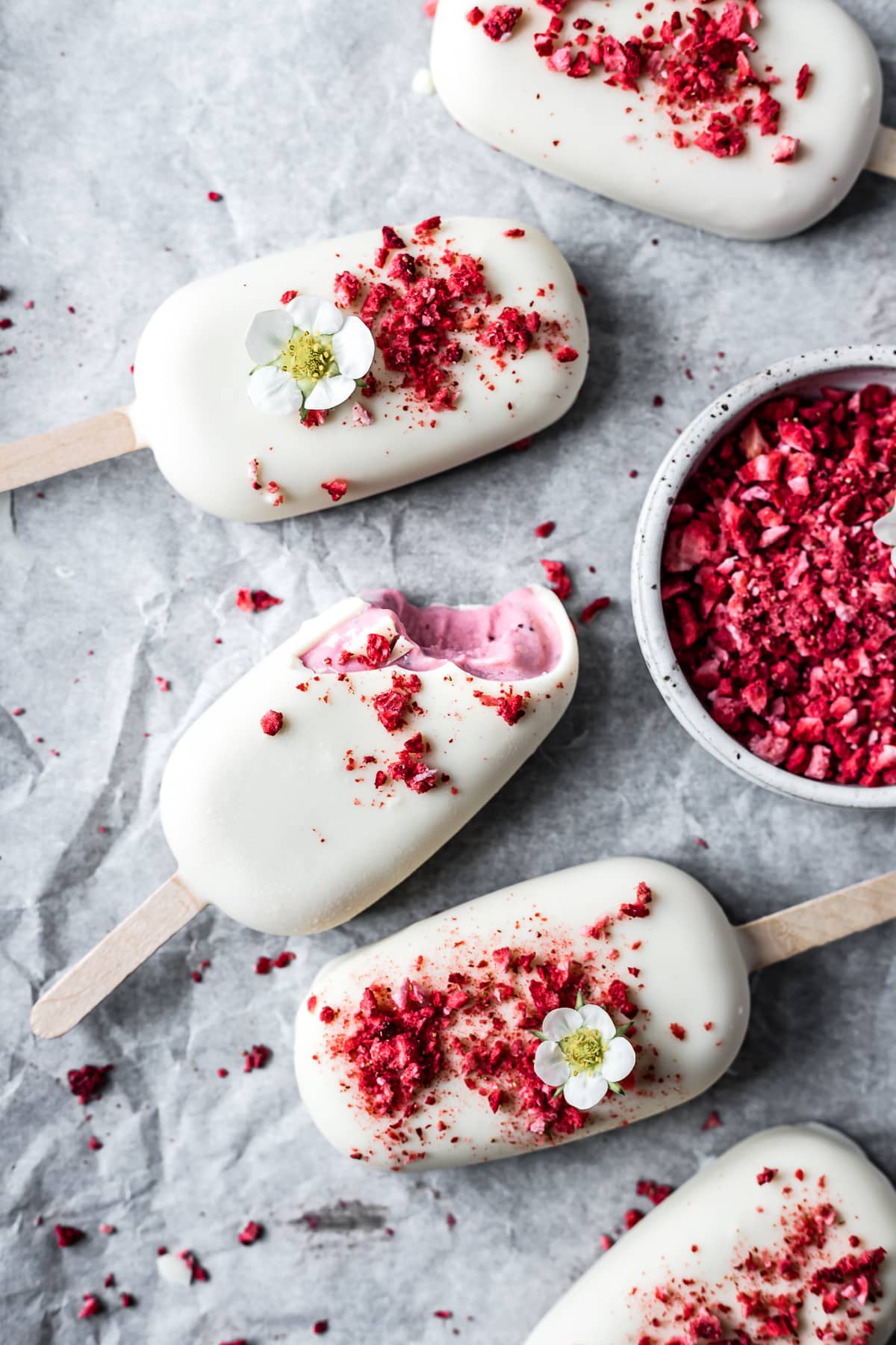 Five ice cream bars on white parchment paper with a bowl of red freeze dried strawberry bits nearby. One of the bars has a bite taken out of it and the pink ice cream is showing. There are strawberry blossoms on two of the bars.