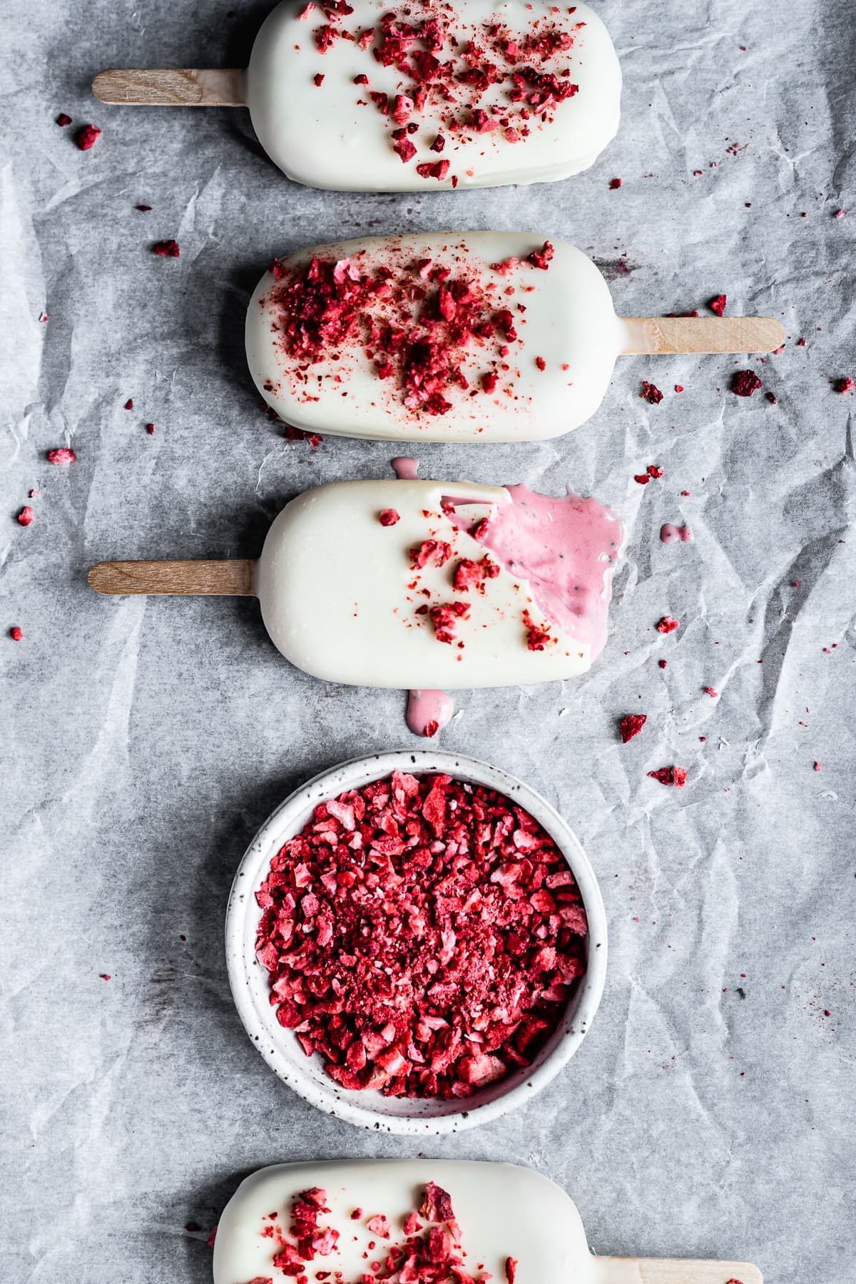 Four ice cream bars in a vertical row with a small bowl of bright red freeze dried strawberries in the middle. All are resting on wrinkled white parchment paper.