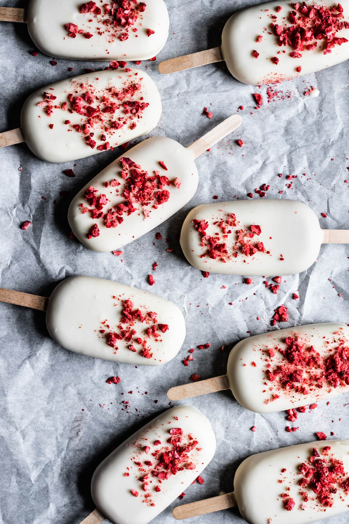 Several white chocolate covered ice cream bars rest on a white parchment paper surface. They have freeze dried strawberries sprinkled onto the white chocolate coating.