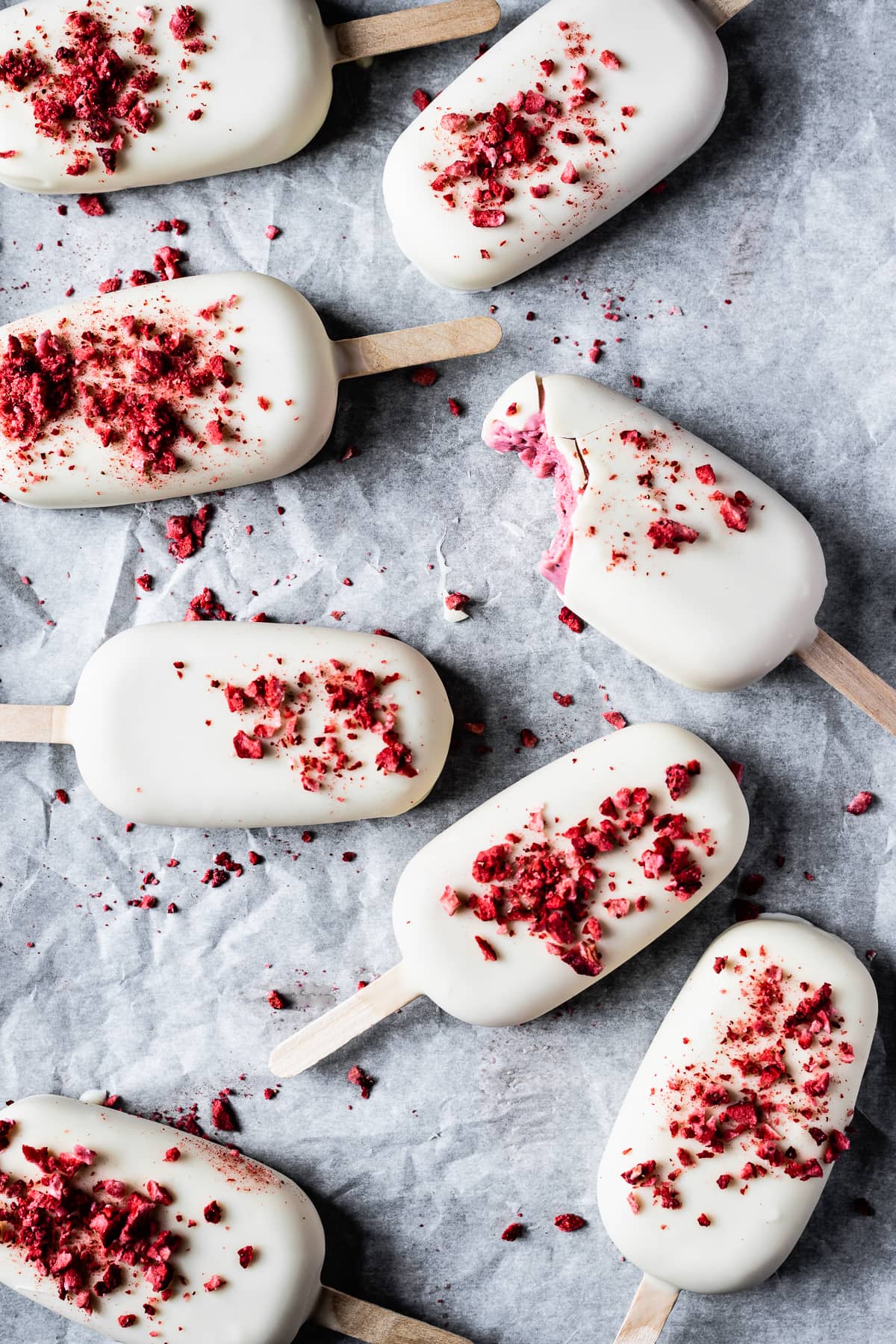 Many white chocolate covered ice cream bars rest on a white parchment paper surface. The main bar has a bite taken out of it, showing the pink ice cream center.