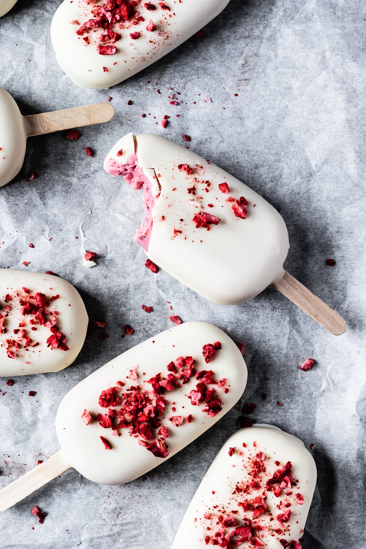 Several white chocolate covered ice cream bars rest on a white parchment paper surface. The main bar has a bite taken out of it, showing the pink ice cream center.