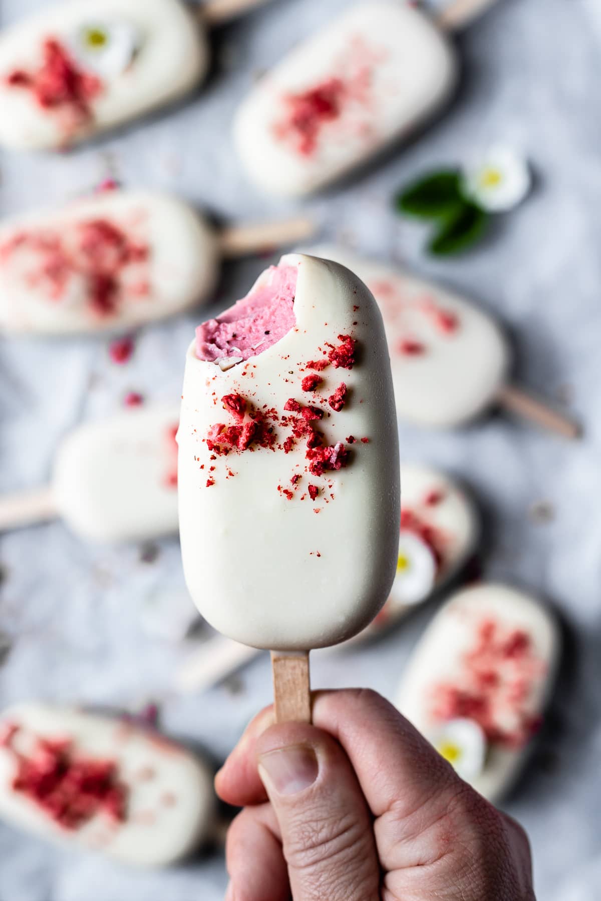 A hand holds an ice cream bar with a bite taken out of it revealing the pink strawberry rhubarb ice cream under the white chocolate shell. There are more ice cream bars in the background.