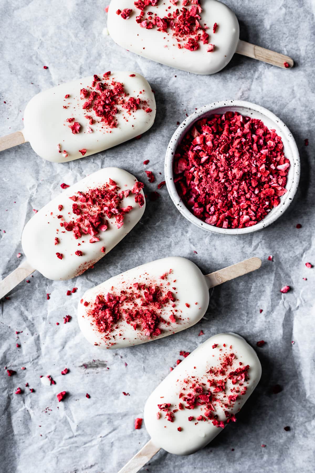 Five ice cream bars on white parchment paper with a bowl of red freeze dried strawberry bits nearby.