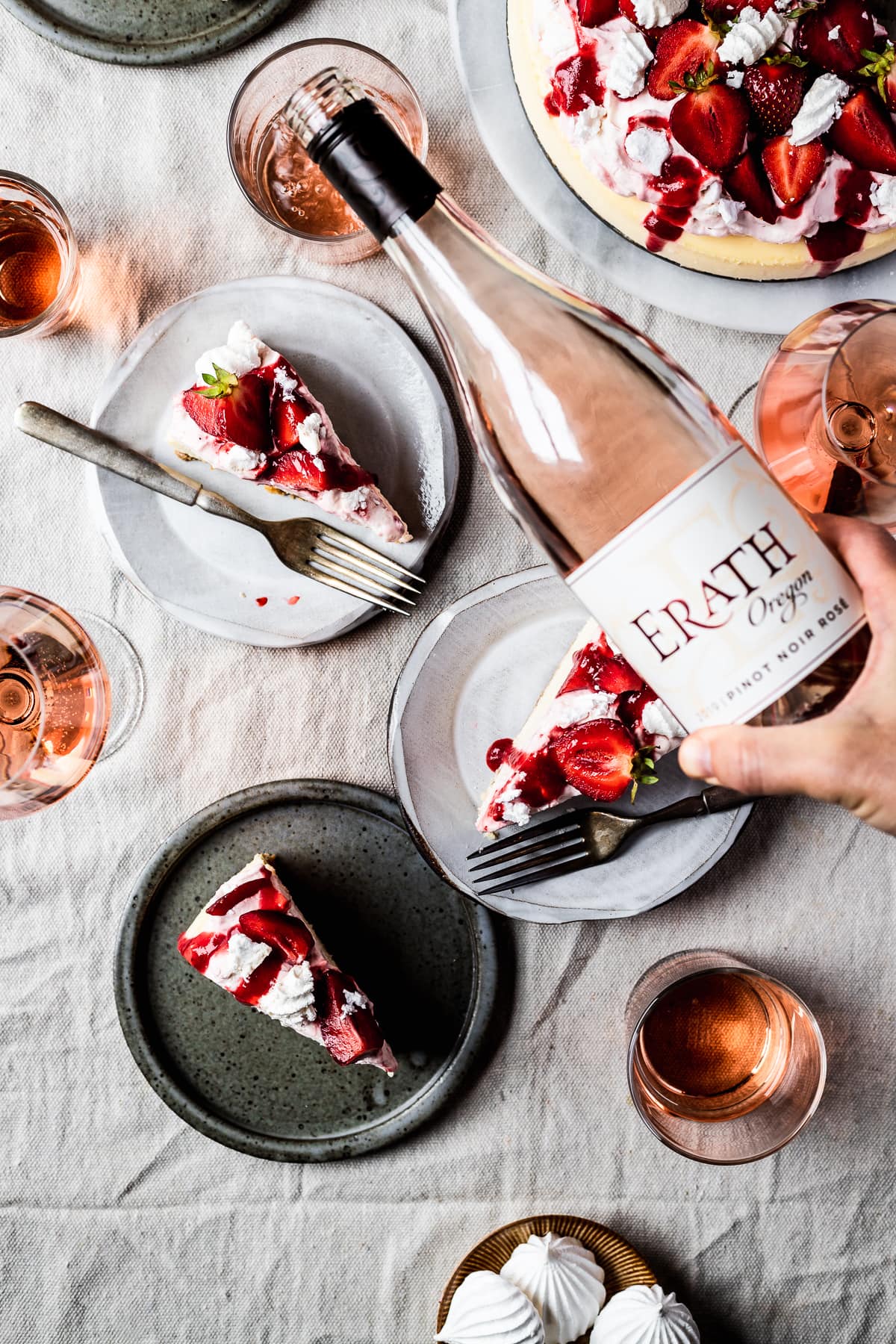 A summery picnic scene with slices of strawberry eton mess cheesecake on plates. A hand pours a bottle of rosé wine into a glass.