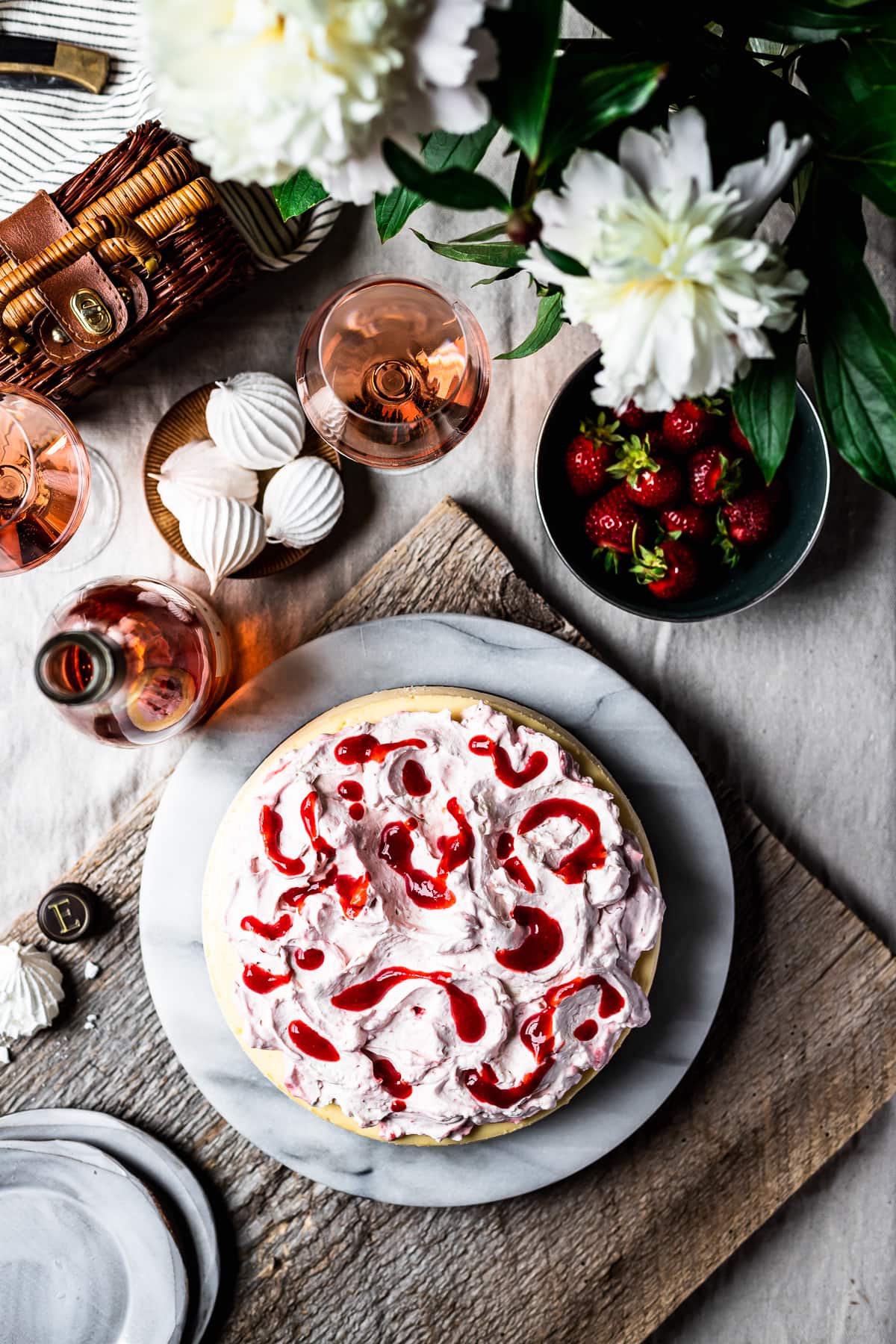 Top view of cheesecake being assembled with whipped cream topping and strawberry coulis. The cheesecake rests on a marble platter on a rustic wooden board. It looks like a picnic scene with a blanket, basket, pocketknife and flowers nearby.