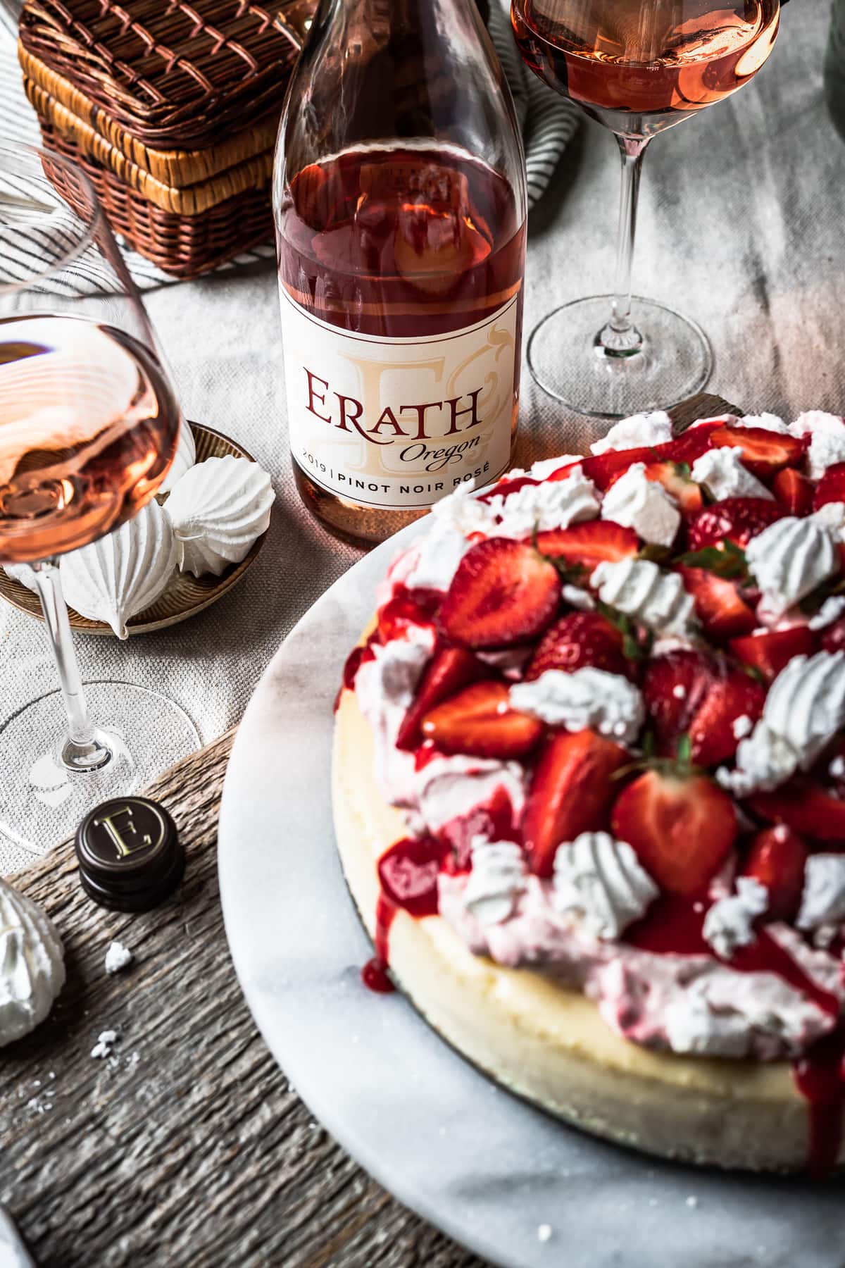 A picnic scene focused a close up of the rosé bottle and the wine in two glasses. An out of focus strawberry cheesecake peeks into the foreground.