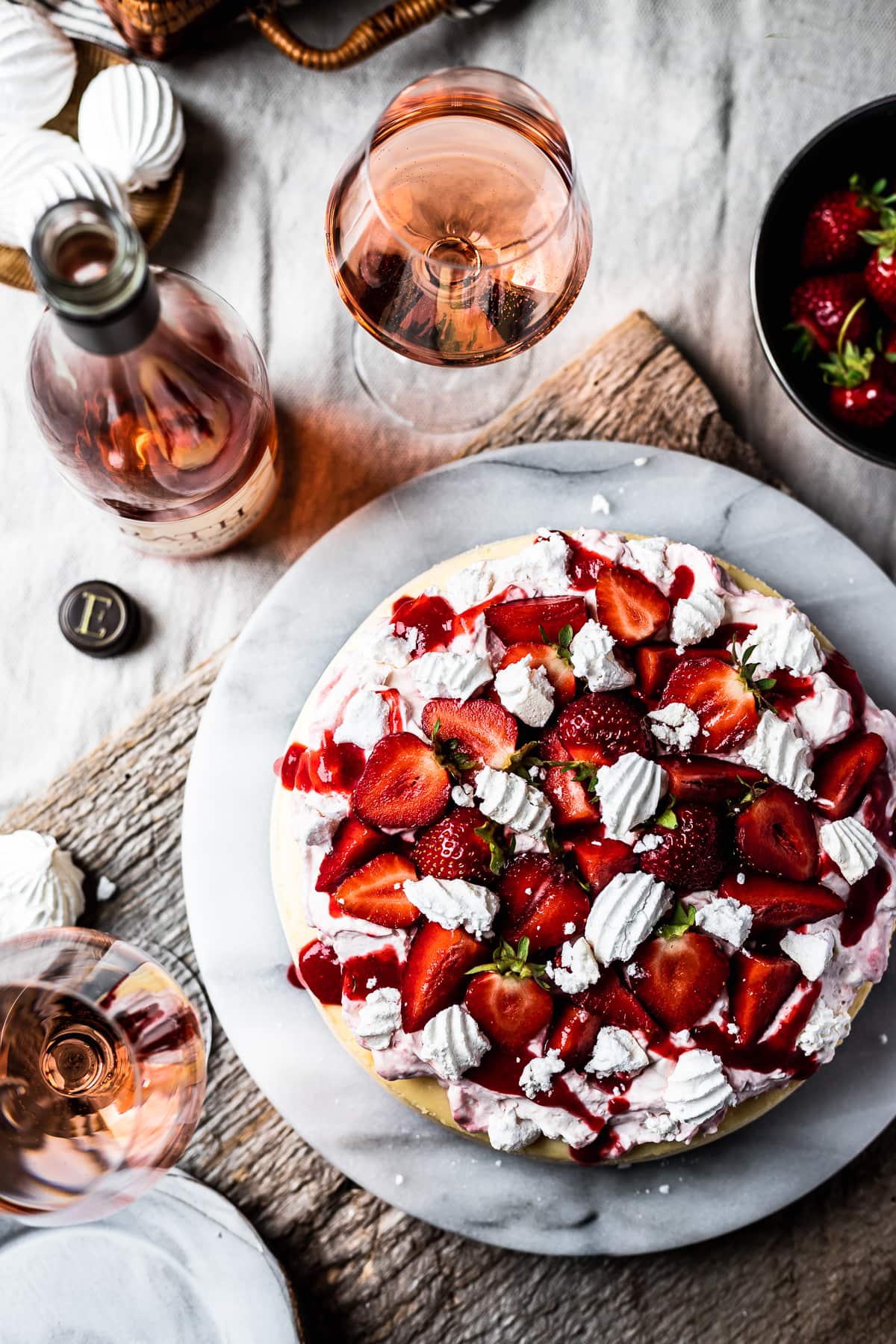 Top view of strawberry topped cheesecake on a marble platter. A rustic wooden board serves as a background. Glasses of rosé wine and a wine bottle rest nearby.