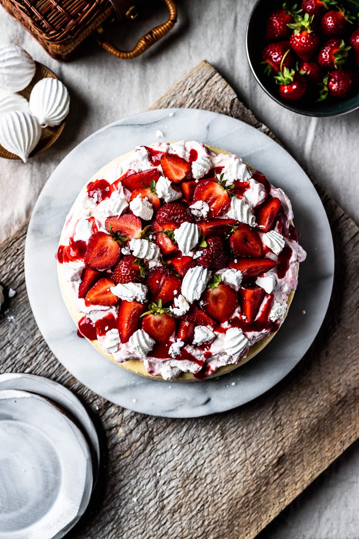 Top view of strawberry topped cheesecake on a marble platter. A rustic wooden board serves as a background. A bowl of fresh strawberries sits nearby.