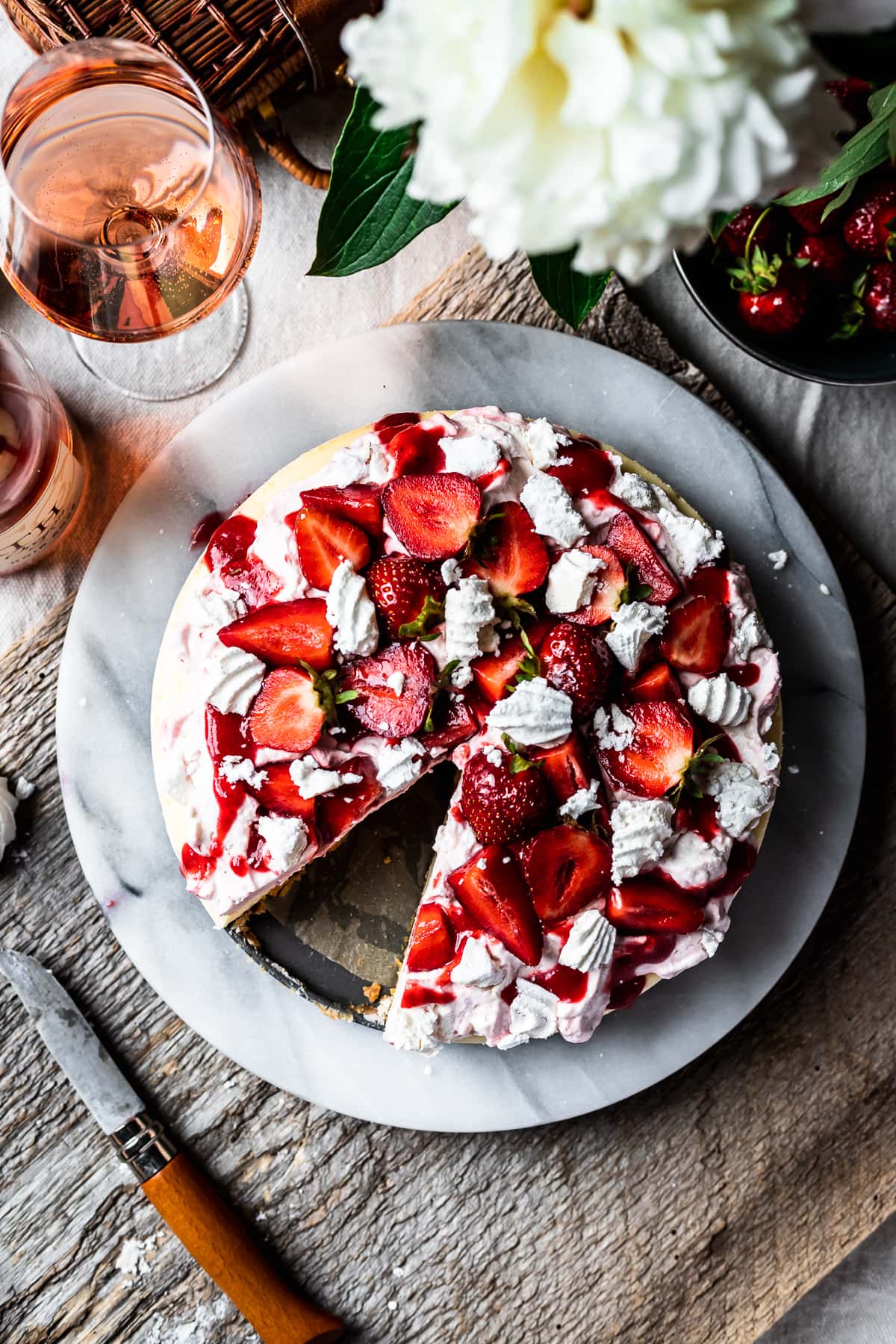 Top view of strawberry topped cheesecake with a slice missing. A rustic wooden board serves as a background. A glass of rosé wine sits nearby.