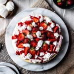 A top view of a vanilla cheesecake topped with whipped cream, strawberry coulis, fresh strawberries and rose water meringues on a marble platter and a rustic wooden cutting board.